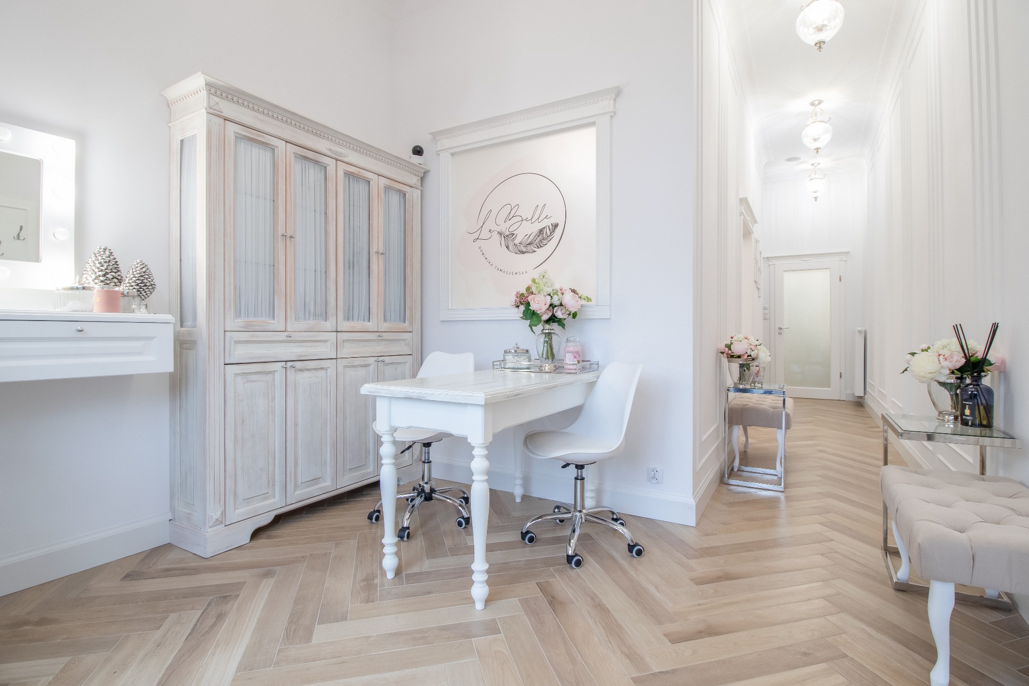 Jasne wnętrze salonu kosmetycznego z drewnianą podłogą w jodełkę, białymi meblami w stylu vintage, kwiatami w wazonach i logo firmy na ścianie.