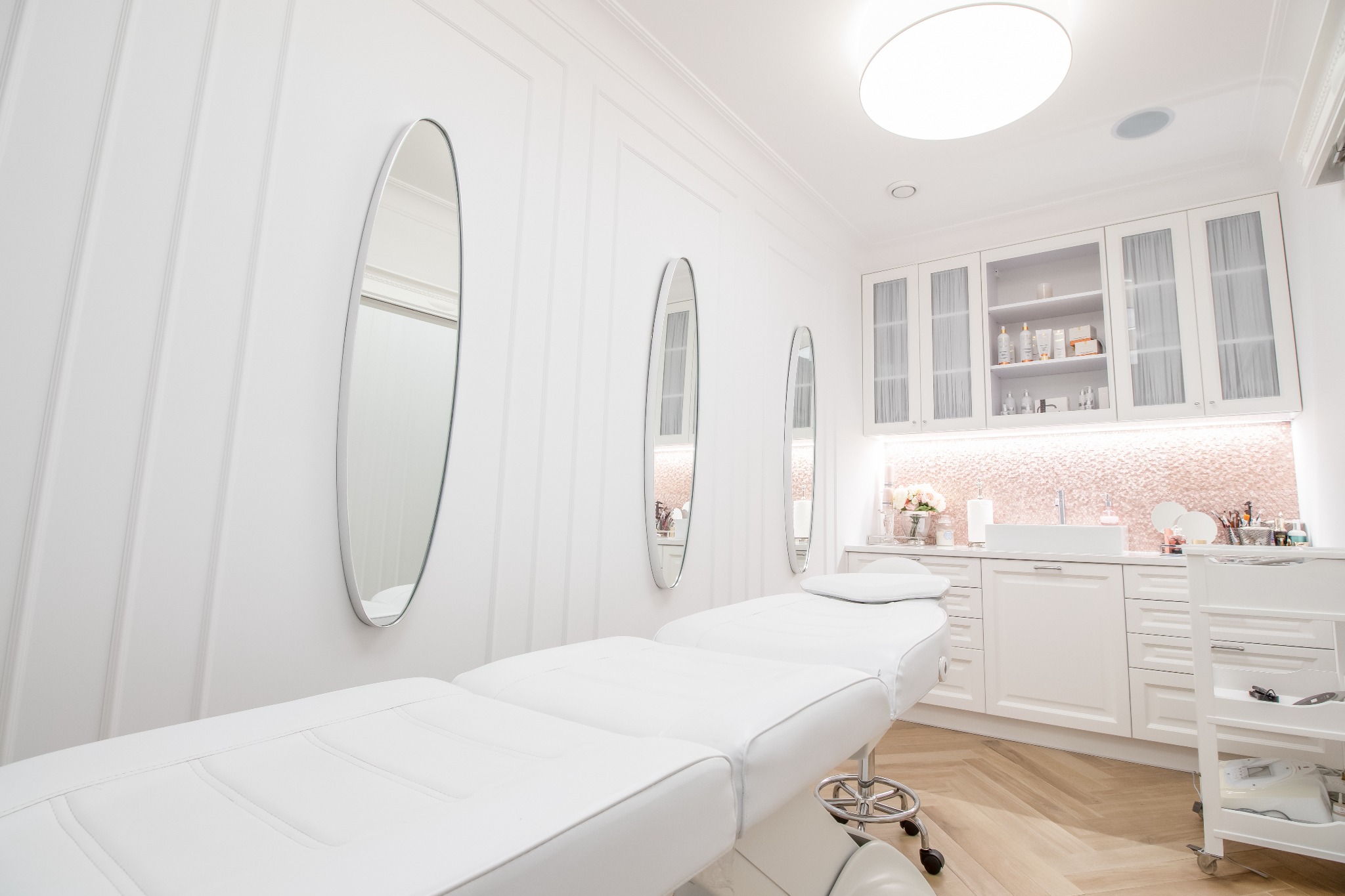 Jasne, minimalistyczne wnętrze gabinetu kosmetycznego z białym leżankiem, trzema owalnymi lustrami na ścianie z panelami oraz zabudową meblową z umywalką i oświetleniem punktowym.