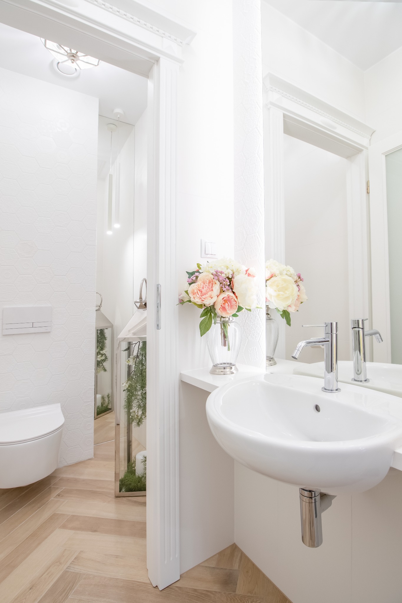 Jasna łazienka z umywalką nablatową, kwiatami w wazonie i podłogą ułożoną w jodełkę. W tle widoczna toaleta i dekoracyjna latarenka z roślinami.