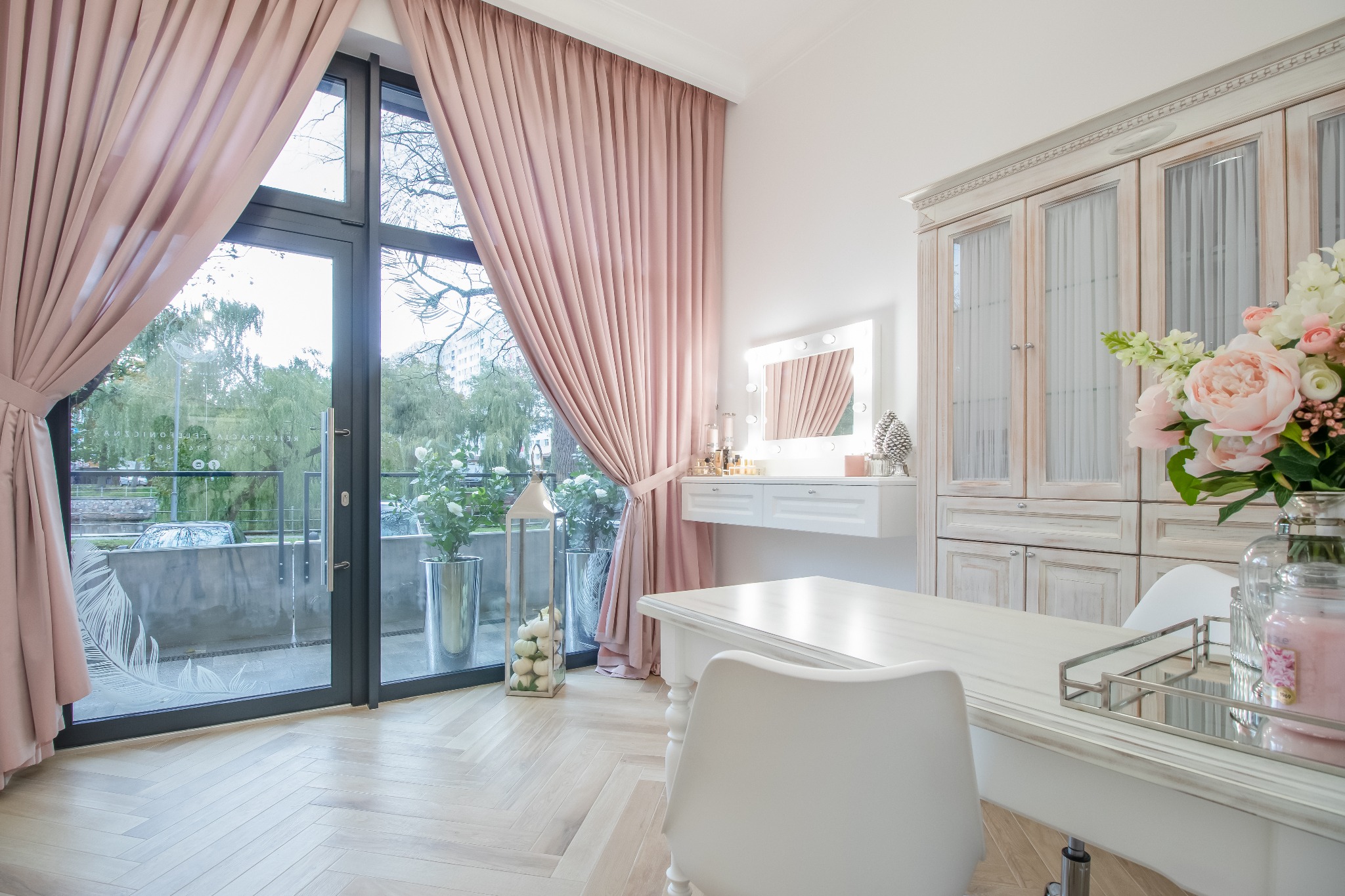 Jasne, przestronne wnętrze pokoju z biurkiem, toaletką z lustrem podświetlanym żarówkami, ozdobną szafką i dużym oknem z różowymi zasłonami, widok na zieleń na zewnątrz.
