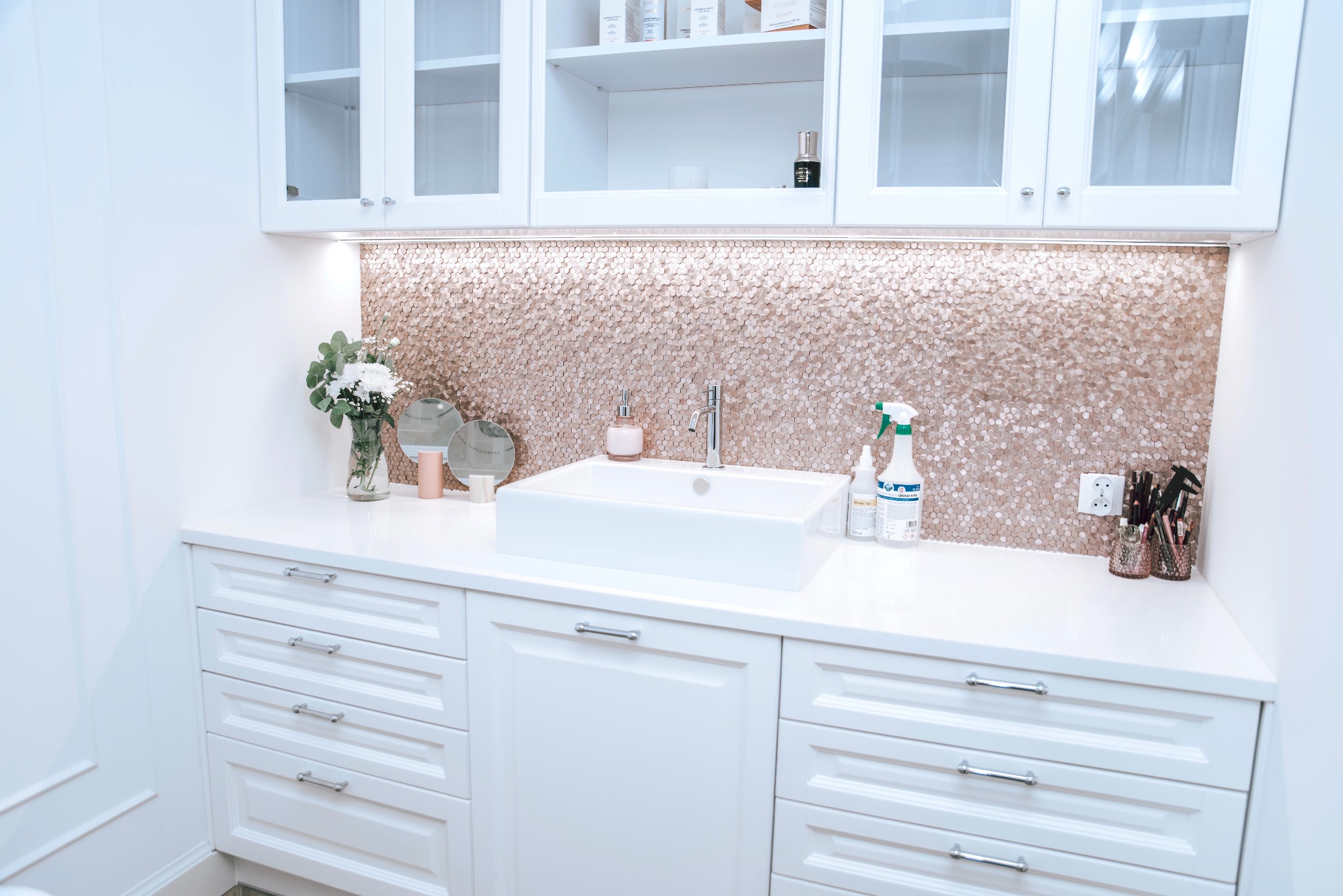 Biała szafka łazienkowa z umywalką nablatową, baterią, mozaiką w odcieniach różowego złota, kwiatami w wazonie i kosmetykami, oświetlona listwą LED pod szafkami wiszącymi.