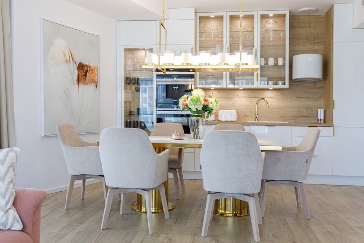Jasna jadalnia z białymi meblami, złotymi akcentami i obrazem pióra na ścianie. Krzesła z beżową tapicerką otaczają stół z marmurowym blatem i złotą podstawą. W tle widoczna zabudowa kuchenna...