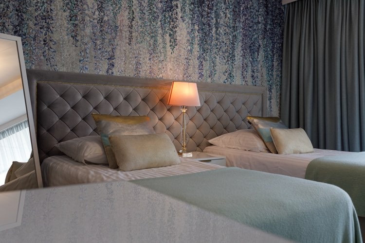 Pokój z dwoma łóżkami z szarym, pikowanym wezgłowiem, lampką nocną z kryształową podstawą i tapetą w kwiaty w tle, widziany z perspektywy lustra.