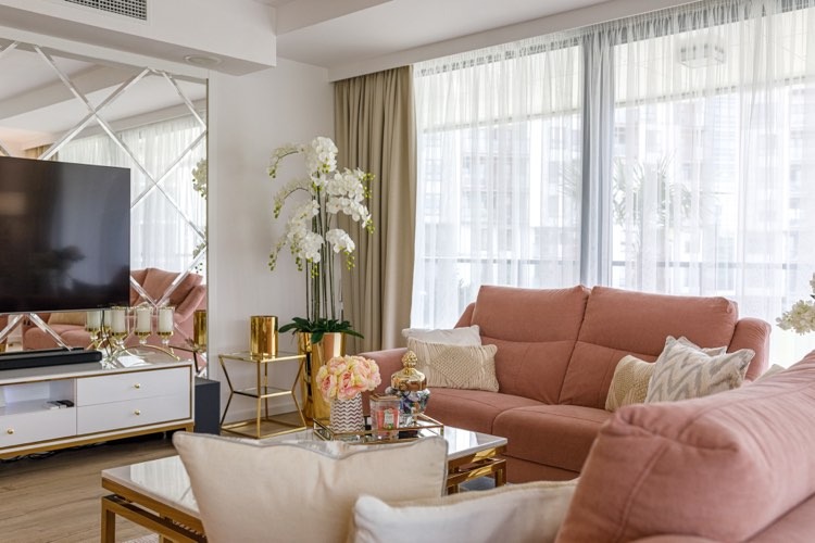 Jasny salon z różową sofą, lustrzaną ścianą z geometrycznym wzorem, białymi meblami i złotymi akcentami dekoracyjnymi, w tym wazonami i stolikami.