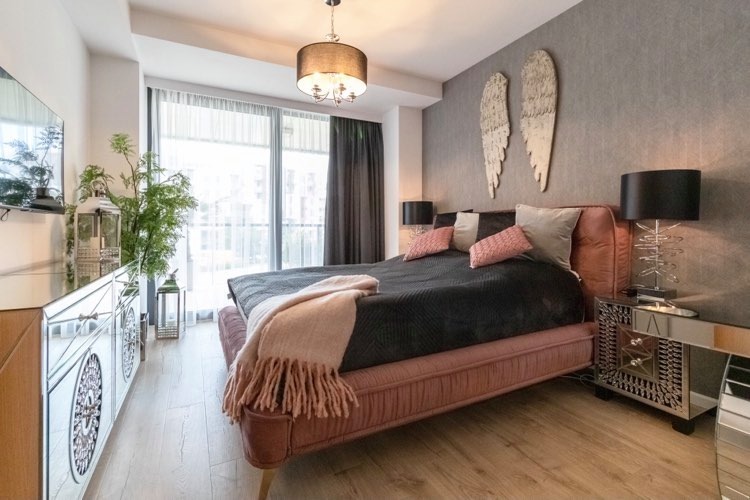 Nowoczesna sypialnia z łóżkiem w odcieniach pudrowego różu i czerni, drewnianą podłogą i dekoracyjnymi skrzydłami na szarej ścianie, oświetlona lampą z abażurem i lampkami nocnymi z metalową podstawą.