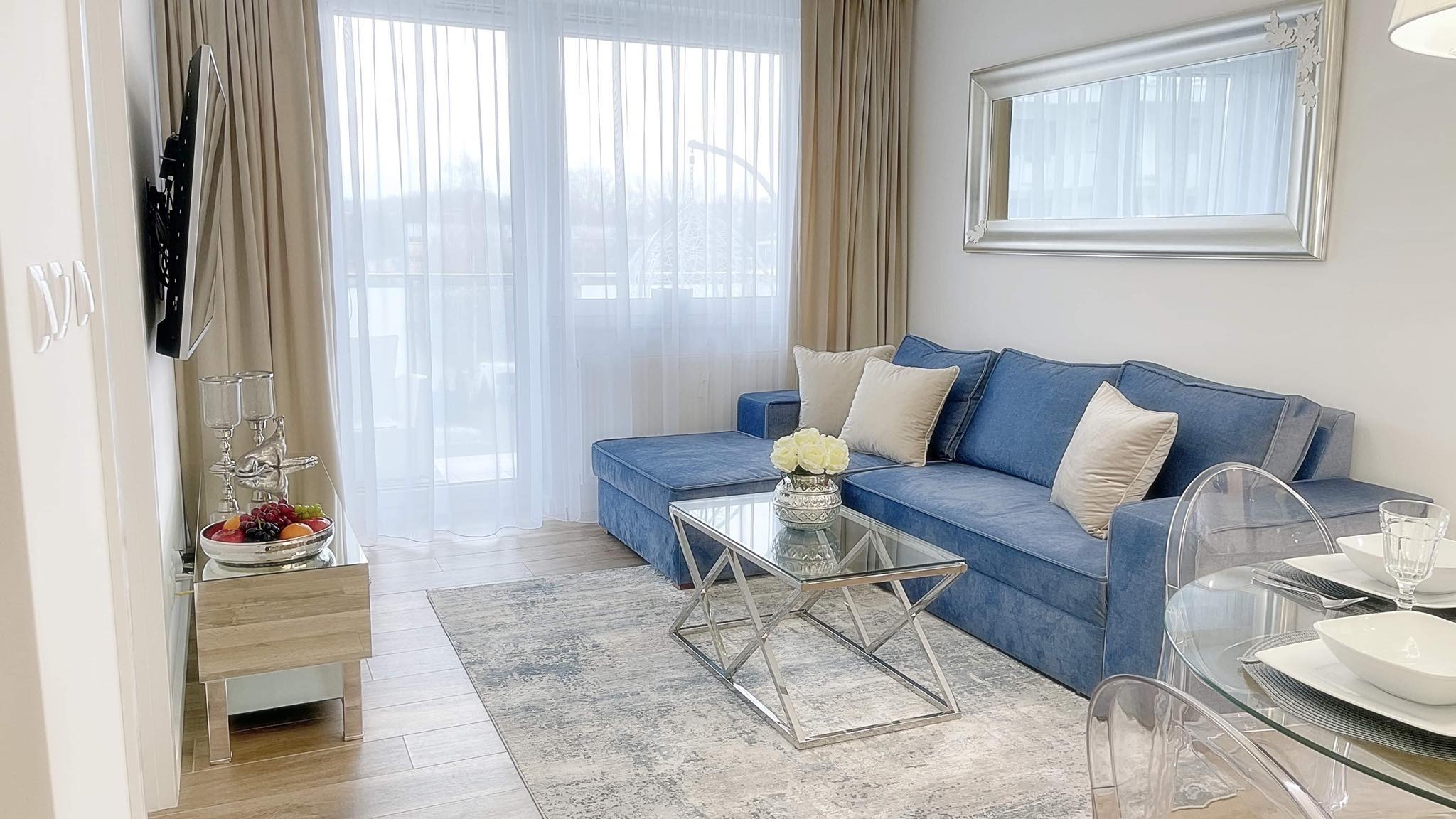 Salon w jasnych barwach z niebieską sofą, szklanym stolikiem kawowym i przezroczystymi krzesłami, dopełniony srebrną ramą lustra i zasłonami w odcieniach beżu.