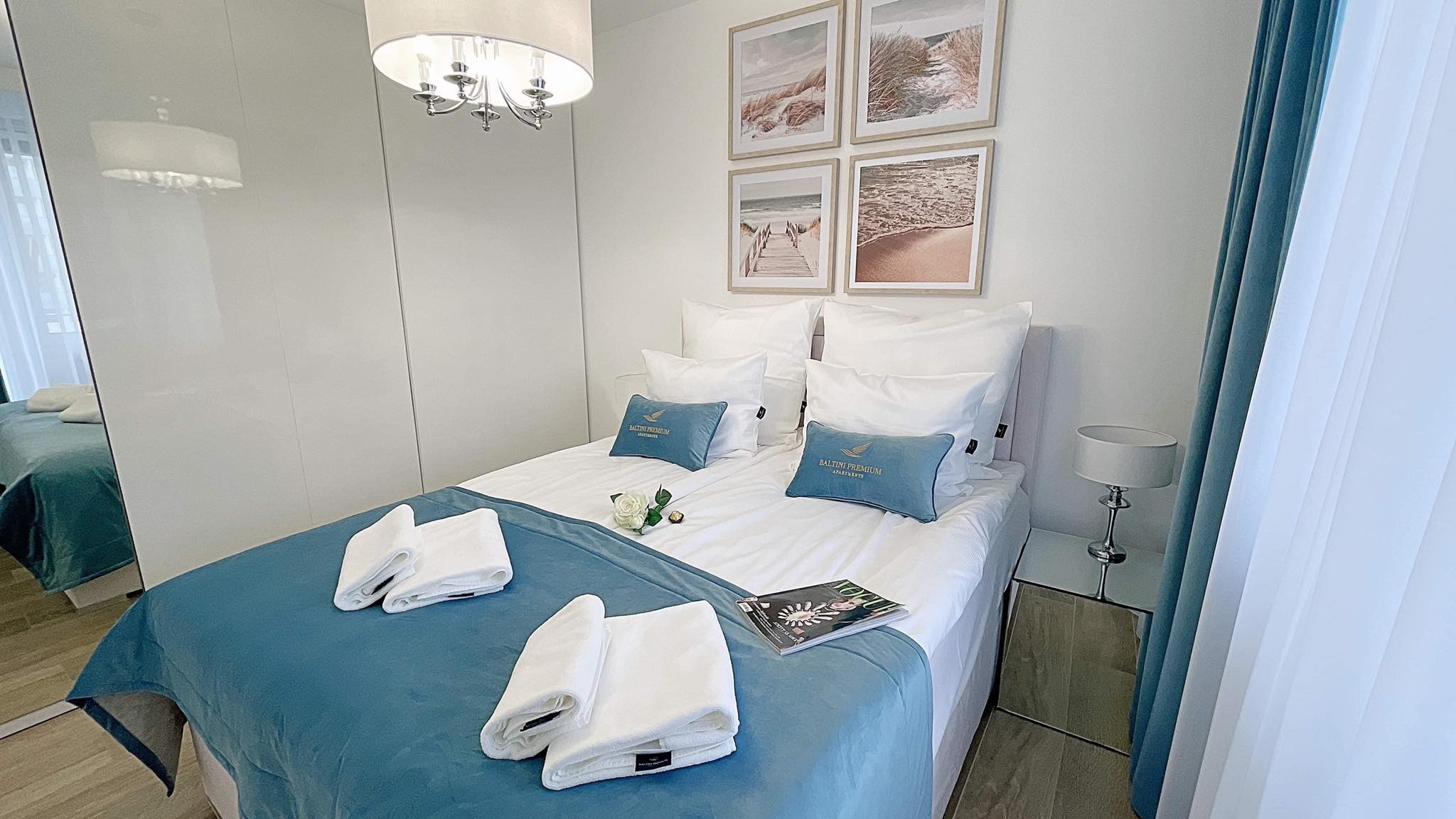 Jasna sypialnia z podwójnym łóżkiem, błękitną narzutą i dekoracyjnymi poduszkami w Kołobrzegu, ściany zdobią obrazy z motywem plaży, a obok łóżka stoi stolik nocny z lampką.