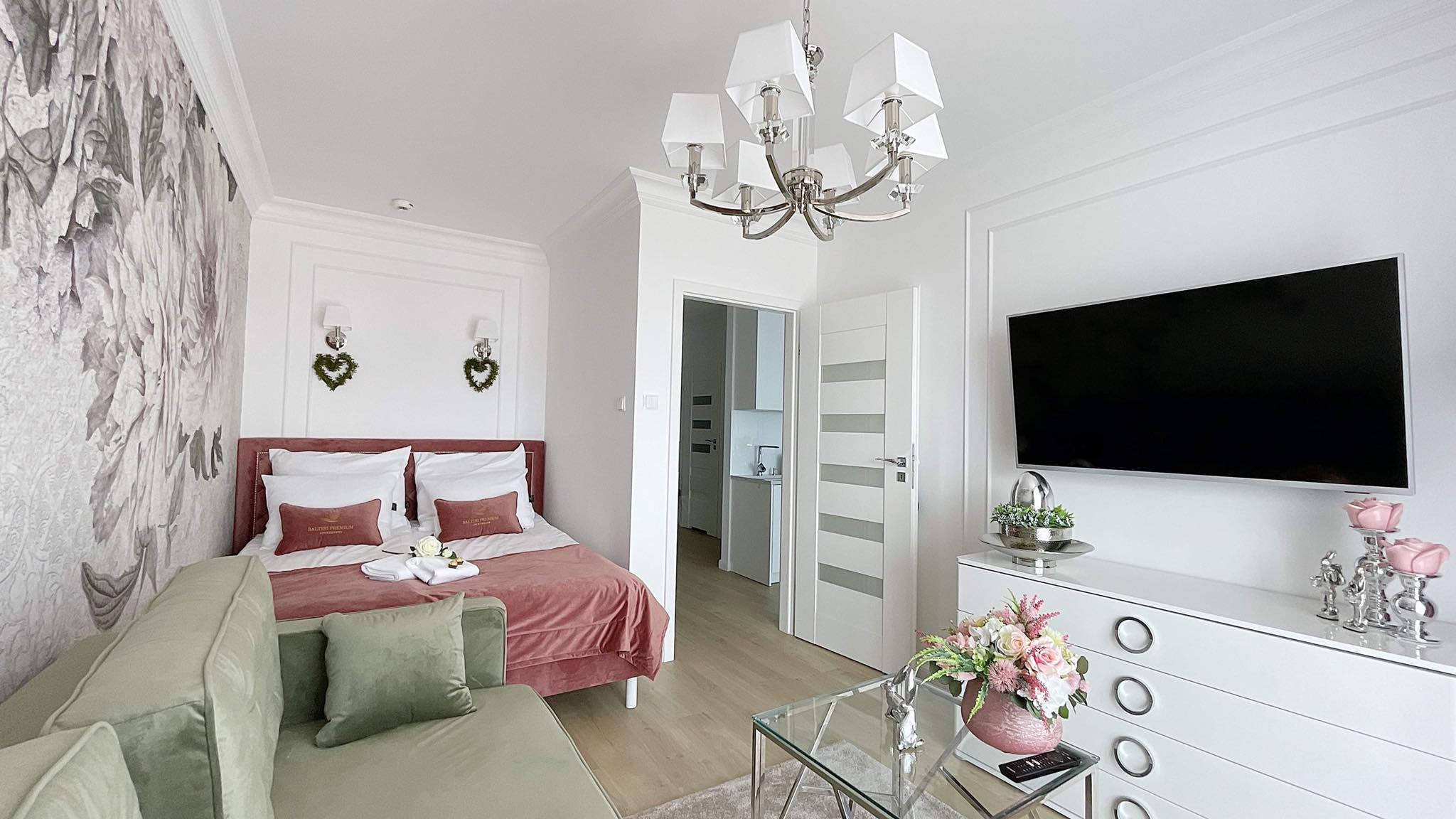 Jasne studio z łóżkiem z różowym wezgłowiem, zieloną sofą, telewizorem na białej komodzie z kwiatami, szklanym stolikiem kawowym i ozdobami w stylu glamour, oświetlone nowoczesnym żyrandolem.