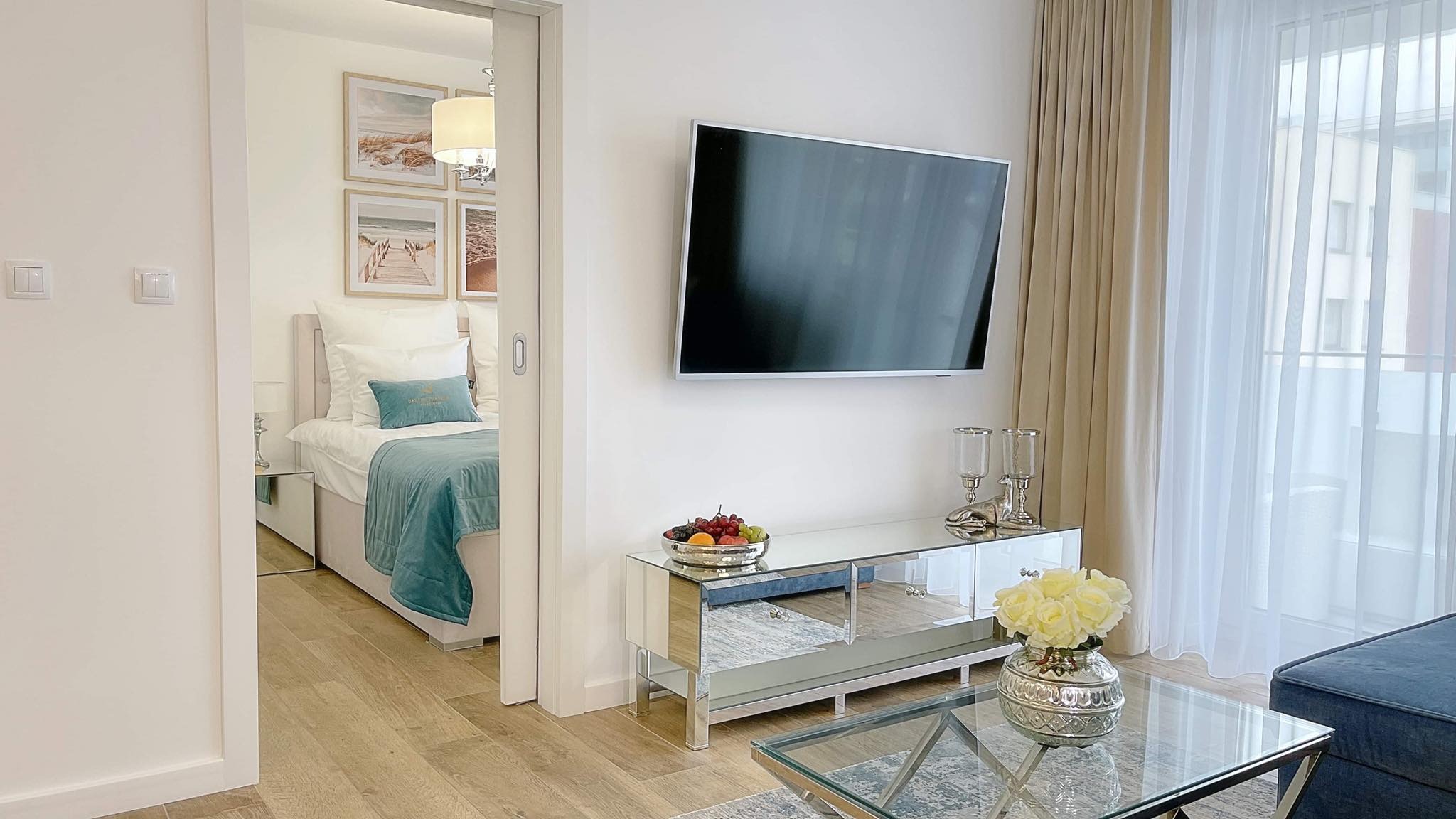 Jasne wnętrze mieszkania z przesuwnymi drzwiami do sypialni, telewizorem na ścianie, lustrzaną komodą z owocami i szklanym stolikiem z kwiatami.