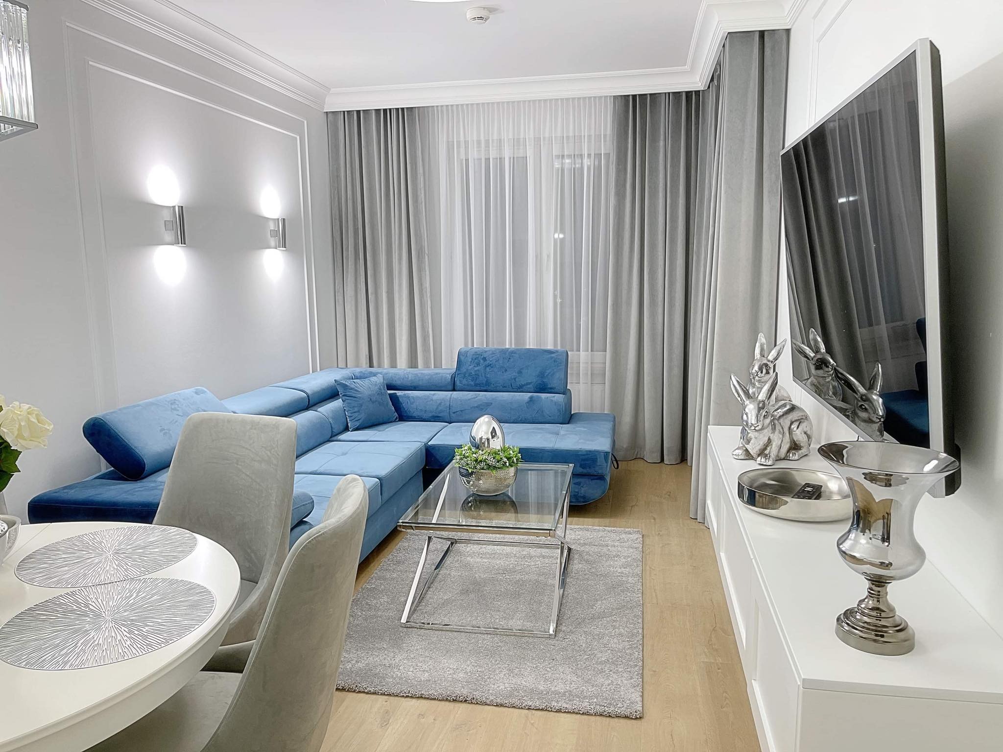 Nowoczesny salon z narożną sofą w odcieniach błękitu, szarym dywanem i stolikiem kawowym ze szklanym blatem, oświetlony srebrnymi kinkietami na ścianie z dekoracyjnymi listwami.