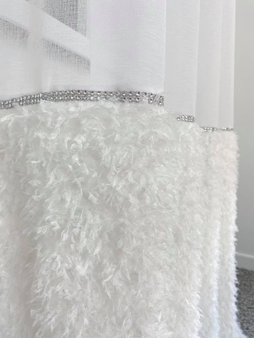 Fragment białej zasłony z dekoracyjnym, futerkowym wykończeniem i srebrnym paskiem z cyrkoniami u góry.