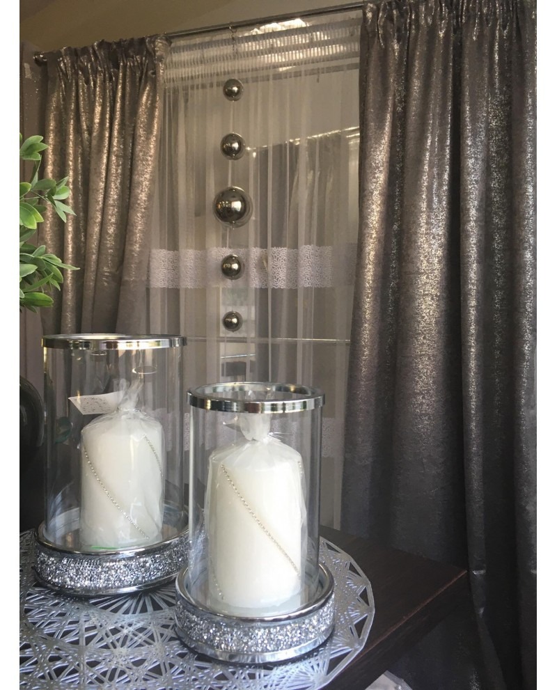 Dekoracyjne, metaliczne zasłony w odcieniach szarości z transparentną warstwą i srebrnymi kulami, uzupełnione dwoma świecami w szklanych lampionach z kryształkami.