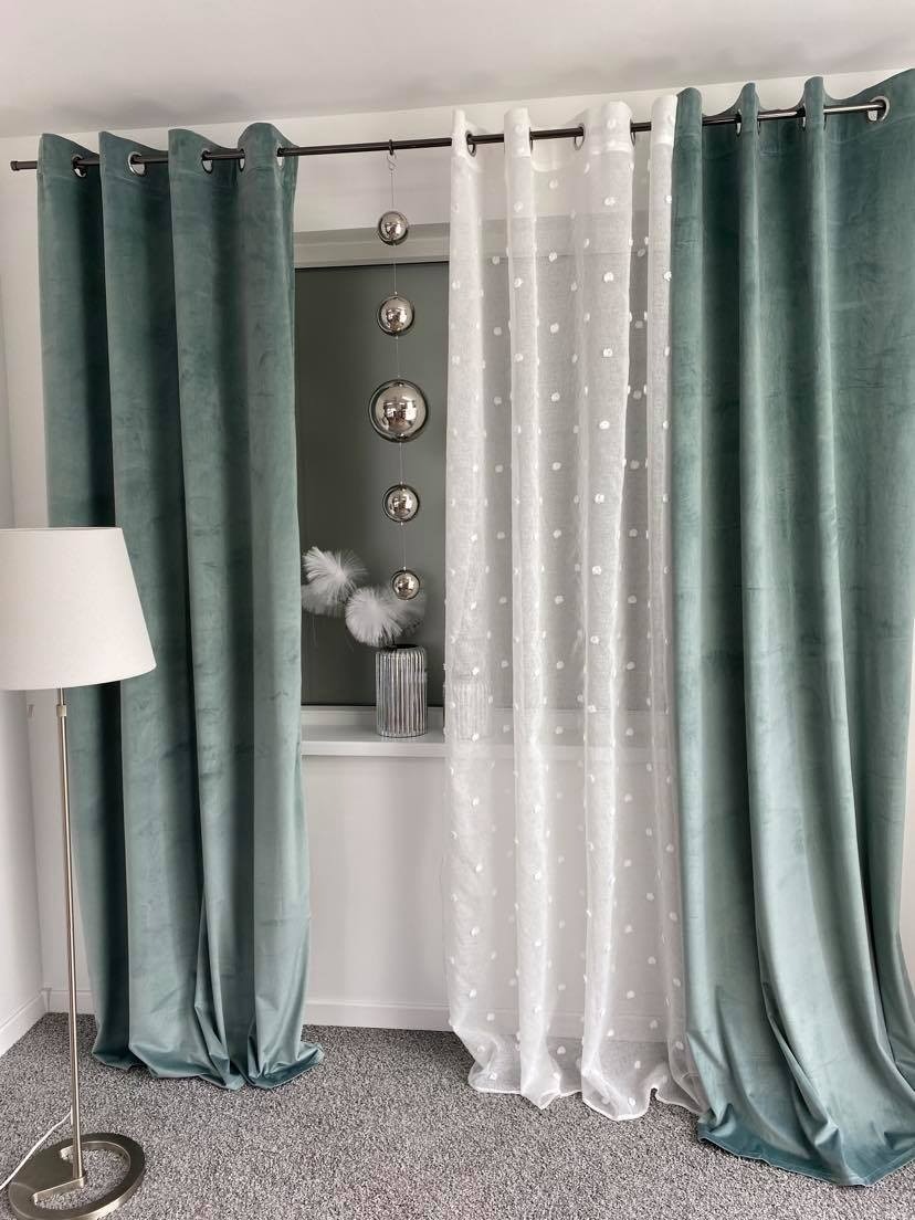 Zestawienie turkusowych welwetowych zasłon z metalowymi oczkami oraz białej firanki w białe kropki, uzupełnione srebrnymi kulami dekoracyjnymi i lampą podłogową