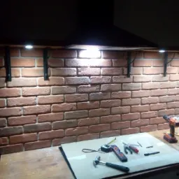 Wykończona ściana z cegły klinkierowej w kuchni z zamontowanymi metalowymi wspornikami i oświetleniem LED, na blacie roboczym narzędzia, w tle czarny okap.