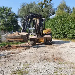 Żółto-czarna koparka Mecalac z łyżką pełną ziemi na żwirowym podłożu, w tle zielone drzewa i krzewy pod błękitnym niebem.