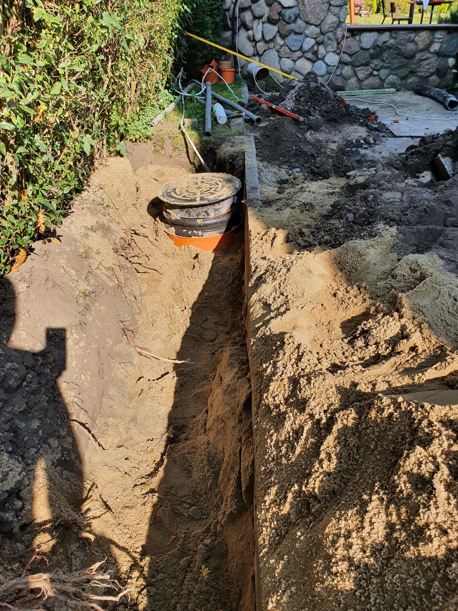 Wykopany rów z widocznym fragmentem zbiornika z czarną pokrywą i pomarańczową podstawą, na tle zieleni i kamiennej ściany.