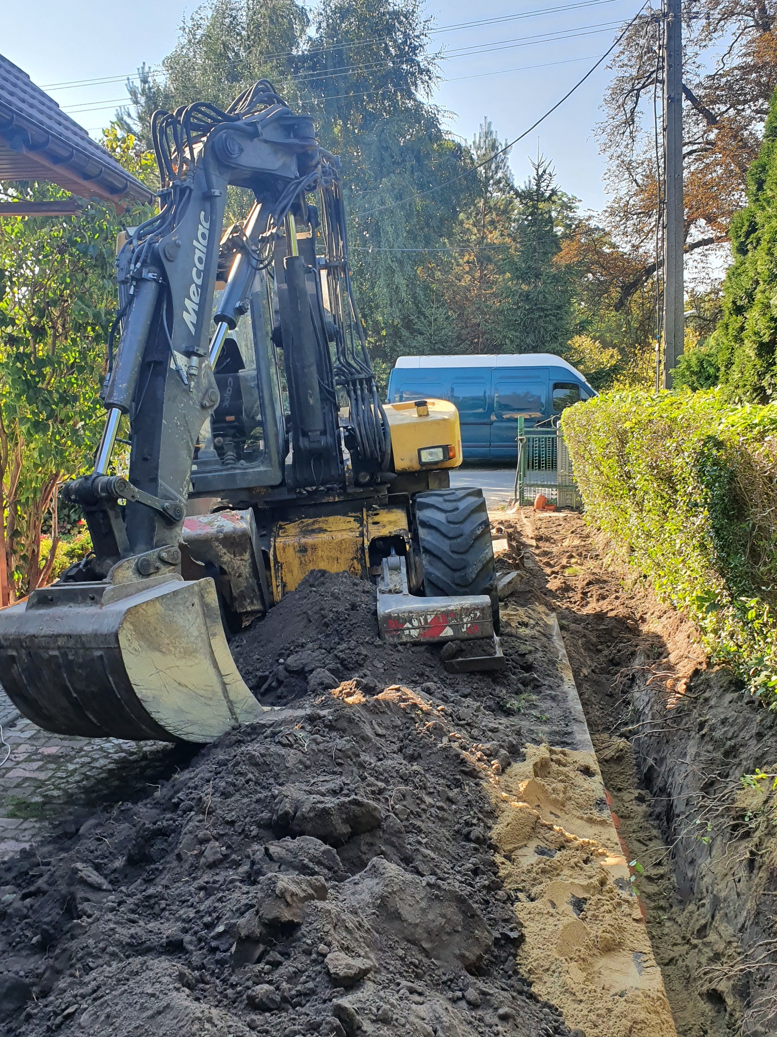 Żółto-czarna koparka Mecalac w trakcie wykonywania wąskiego wykopu w ziemi, obok żywopłotu i chodnika z kostki, z widoczną niebieską furgonetką w tle.