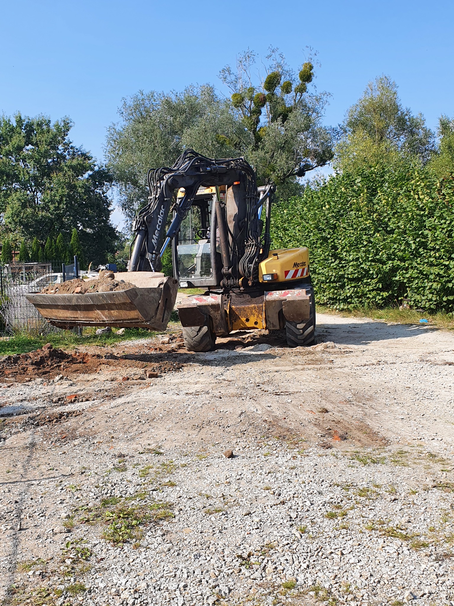 Żółto-czarna koparka Mecalac z łyżką pełną ziemi na żwirowym podłożu, w tle zielone drzewa i krzewy pod błękitnym niebem.