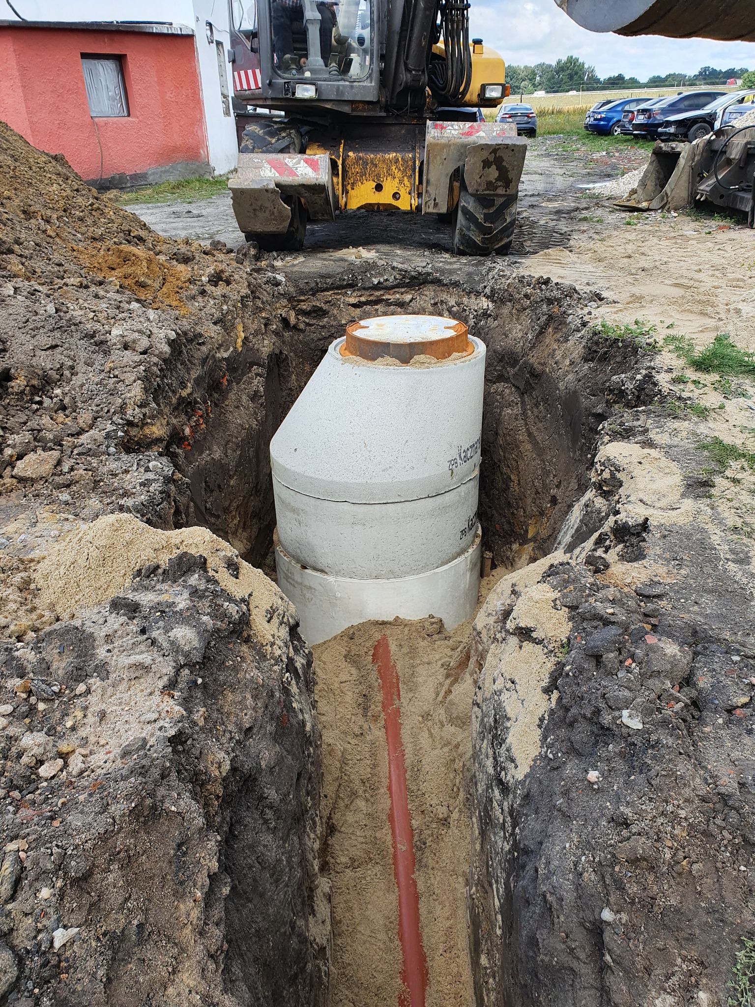 Betonowy zbiornik, prawdopodobnie szambo, umieszczony w wykopie, widoczny rurociąg, w tle koparka i budynek.