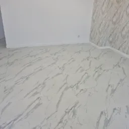 Wykończone wnętrze pokoju z białymi ścianami, podłogą z płytek imitujących biały marmur z szarymi żyłami oraz fragmentem ściany wykończonej tapetą w podobnym wzorze.