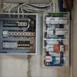 Dwie skrzynki elektryczne z otwartą obudową po lewej stronie, widoczne liczne kable i bezpieczniki, zamontowane na surowej, betonowej ścianie. Nad nimi widoczne rury osłonowe z kablami.