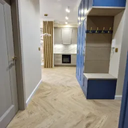 Nowoczesne wnętrze: korytarz z szafą w zabudowie w odcieniach granatu i beżu, parkiet ułożony w jodełkę, widok na szarą kuchnię z białym blatem i marmurowym backsplash