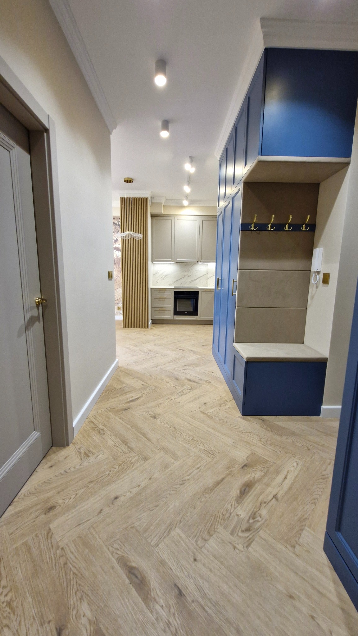 Nowoczesne wnętrze: korytarz z szafą w zabudowie w odcieniach granatu i beżu, parkiet ułożony w jodełkę, widok na szarą kuchnię z białym blatem i marmurowym backsplash
