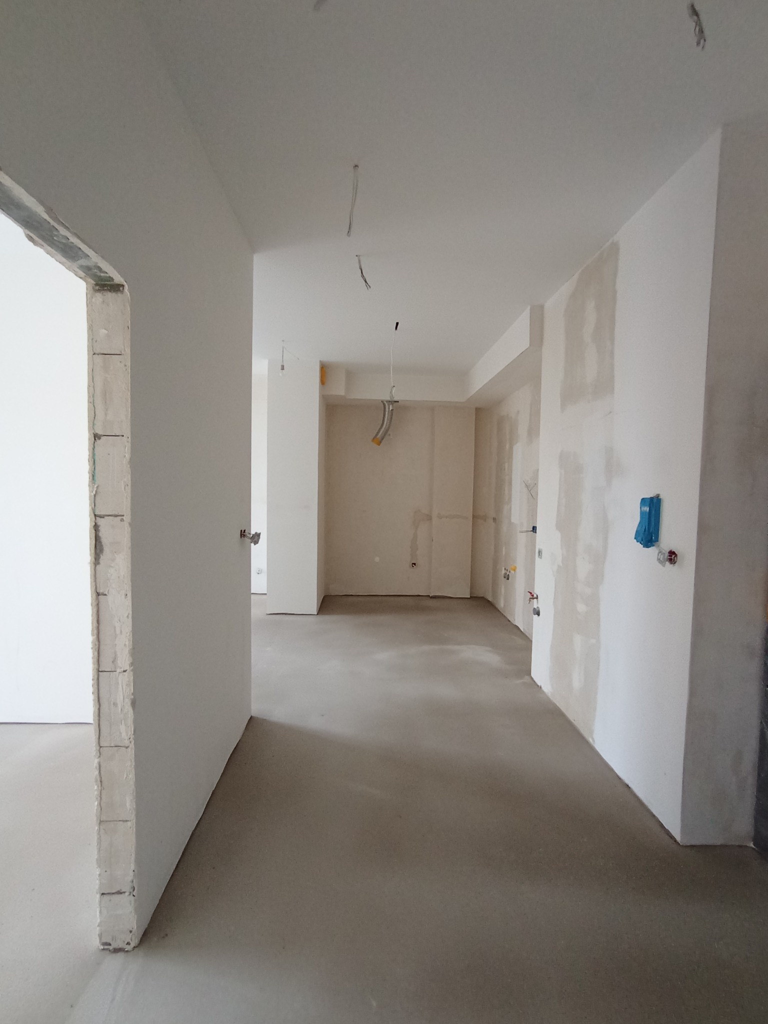 Wnętrze w trakcie prac wykończeniowych: gładkie, szare wylewki podłogowe, ściany częściowo pokryte gładzią, widoczne instalacje elektryczne, surowy otwór drzwiowy.