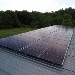 Panele słoneczne zamontowane na blaszanym dachu, widok z góry, w tle zieleń drzew i pochmurne niebo.