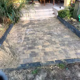 Świeżo ułożona, brązowo-szara kostka brukowa z czarnym obrzeżem, częściowo zasypana piaskiem, prowadząca do schodów tarasu. Z boku widoczna zieleń i zielona konewka.