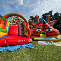 Dwa kolorowe dmuchane zamki na trawie: jeden z motywem dżungli i zjeżdżalnią, drugi w kształcie żyrafy z dużymi oczami, na tle budynku i drzew.