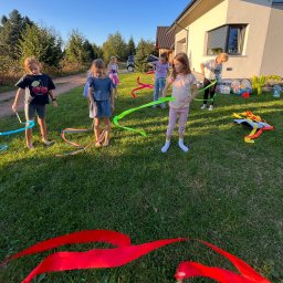 Szalone Figle - Grupa dzieci na trawie bawi się kolorowymi wstążkami, w tle dom i drzewa, widać akcesoria do zabawy.