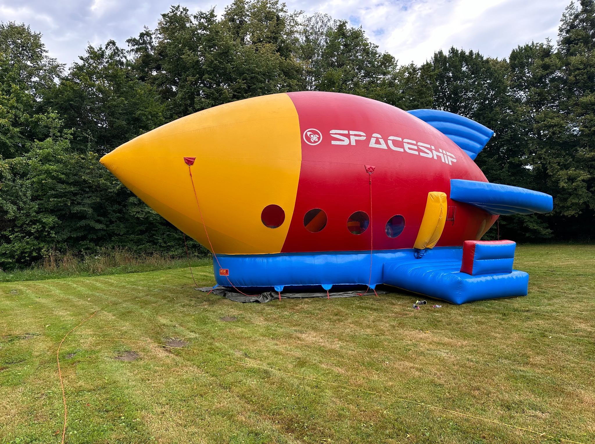 Nadmuchiwany statek kosmiczny w kolorach żółtym, czerwonym i niebieskim stoi na trawniku przed drzewami. Widoczne wejścia do dmuchańca i logo 'Spaceship'.