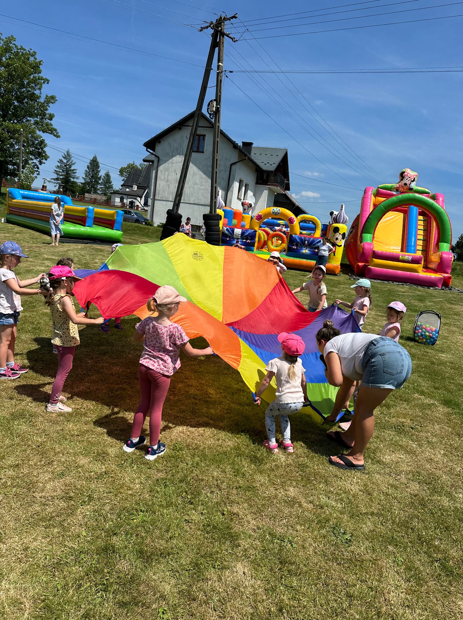 Grupa dzieci i dorosły bawią się kolorową chustą animacyjną na trawie, w tle dmuchane zamki i dom z liniami energetycznymi.