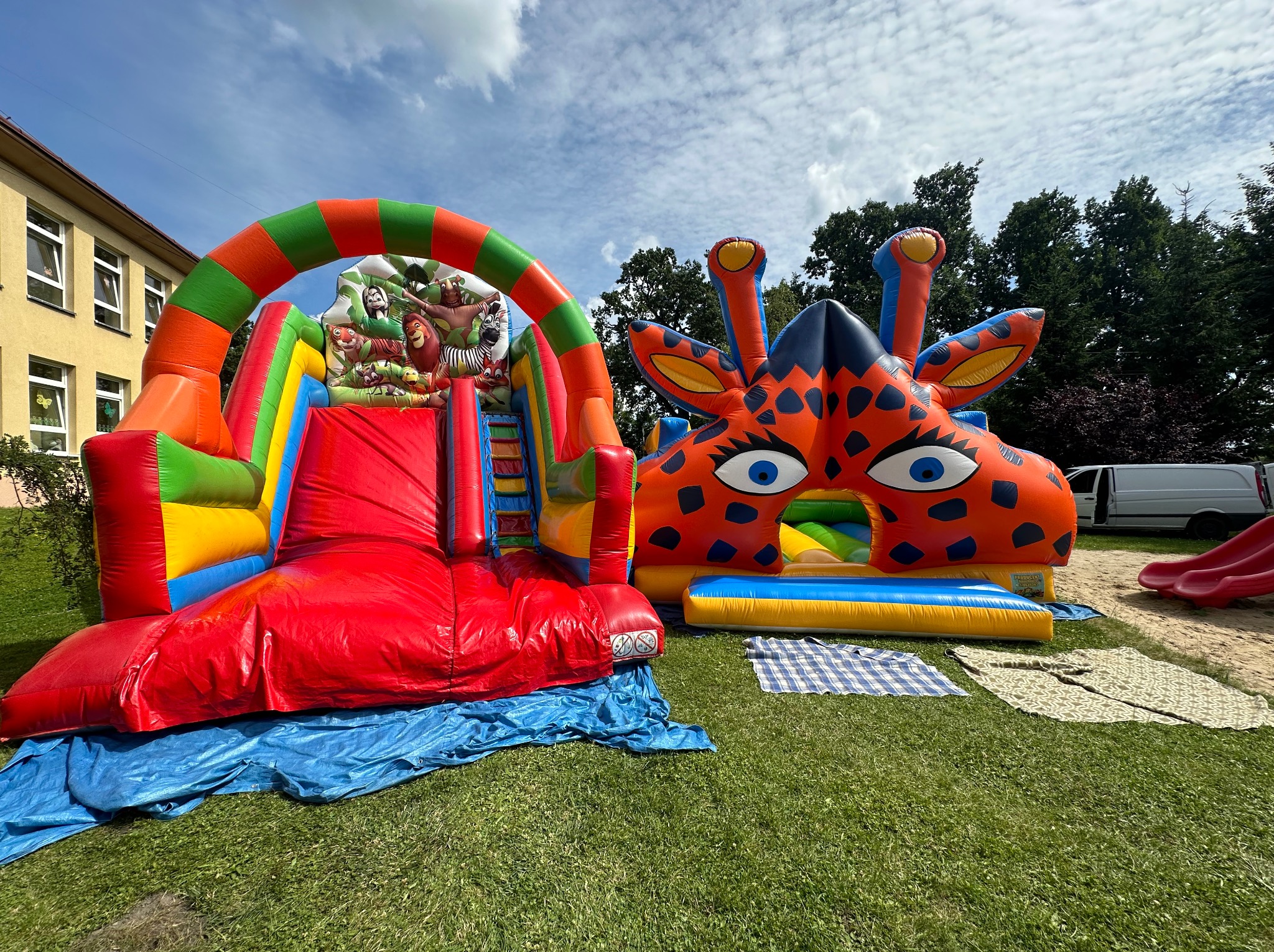 Dwa kolorowe dmuchane zamki na trawie: jeden z motywem dżungli i zjeżdżalnią, drugi w kształcie żyrafy z dużymi oczami, na tle budynku i drzew.