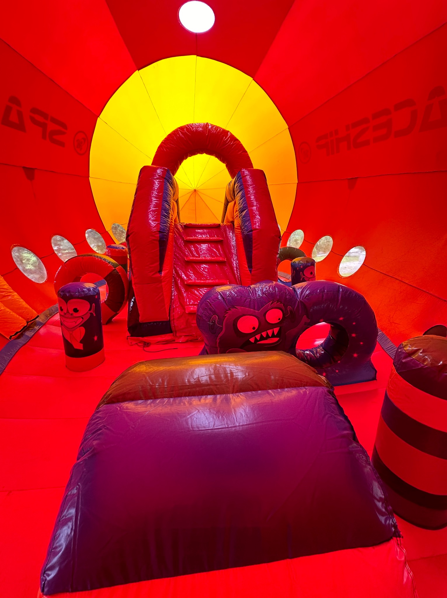 Wnętrze czerwonego dmuchanego zamku z żółtym oknem w suficie, dmuchana zjeżdżalnia i przeszkody w kształcie potworów.