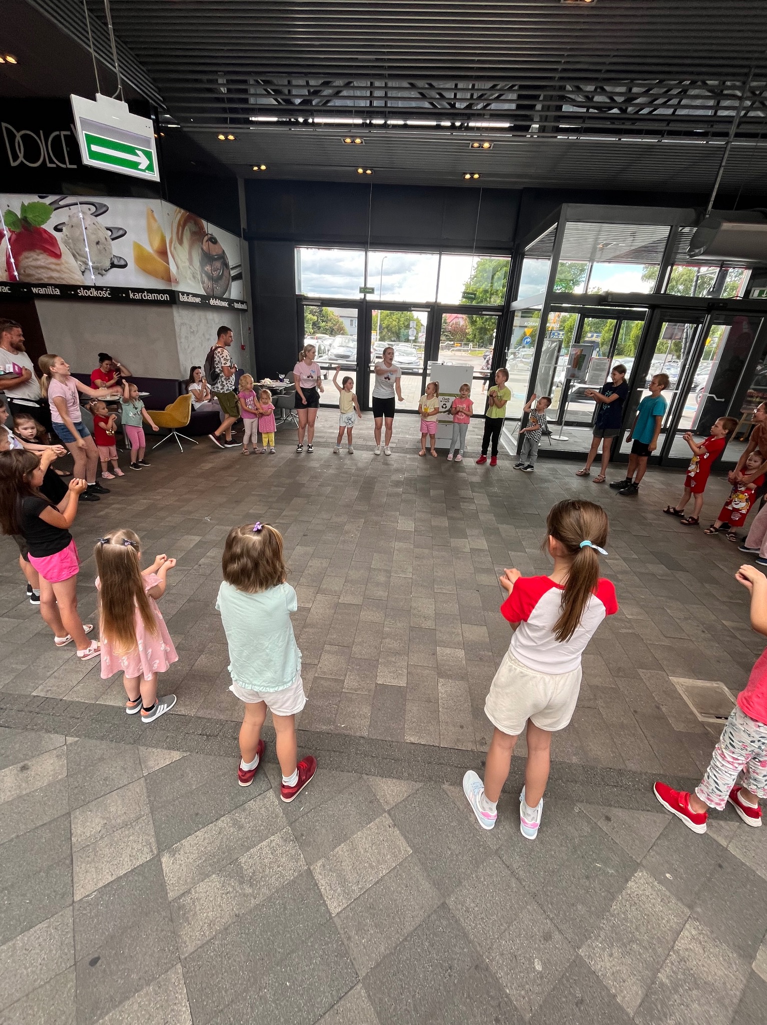Grupa dzieci ustawiona w kręgu, prawdopodobnie podczas zabawy lub zajęć z animatorem w przestrzeni publicznej, z dorosłymi obserwującymi w tle.