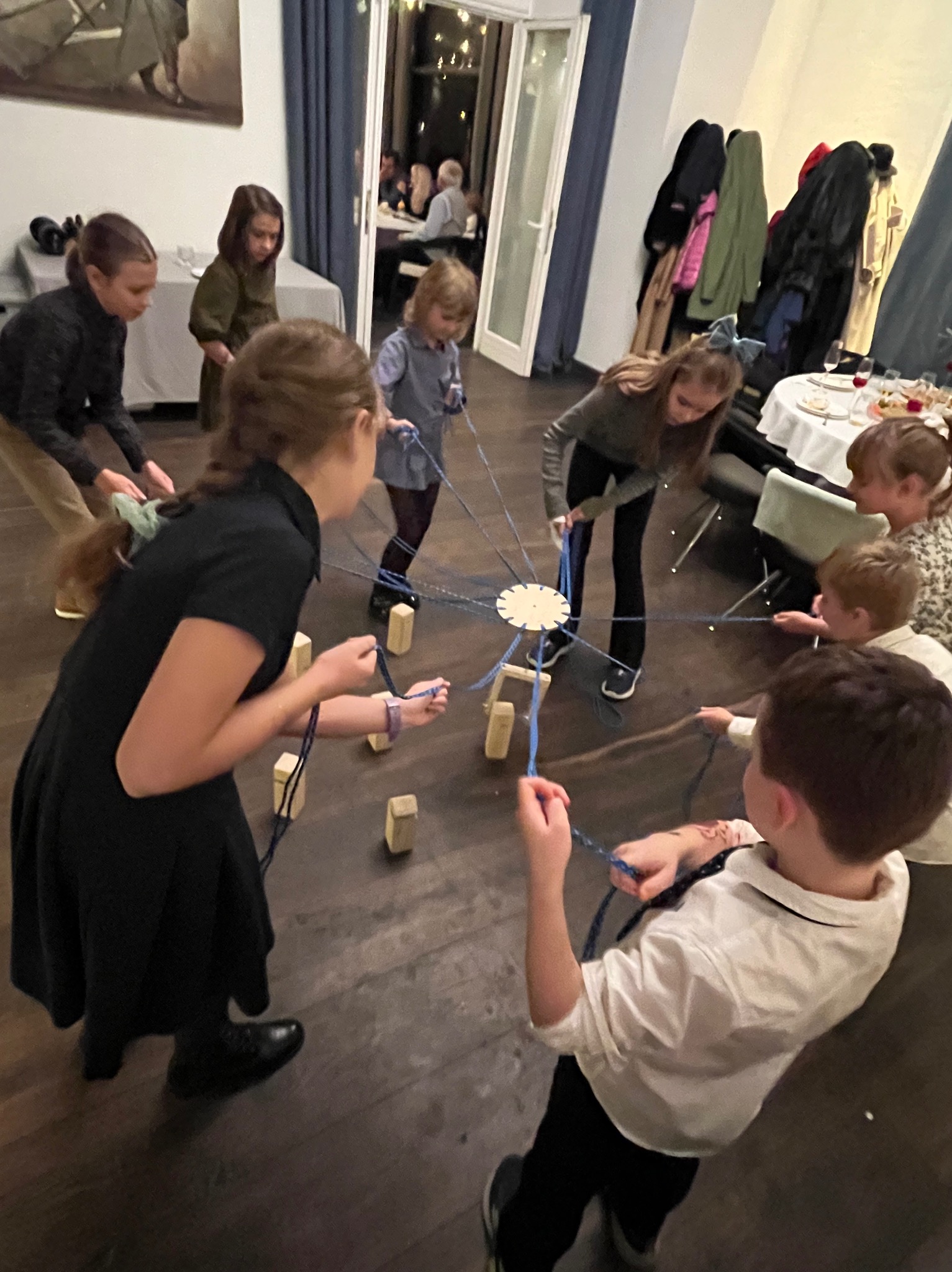 Grupa dzieci w różnym wieku, w otoczeniu drewnianych klocków i sznurków, zaangażowana w interaktywną zabawę w pomieszczeniu z parkietem, w tle widoczni dorośli siedzący przy stole.