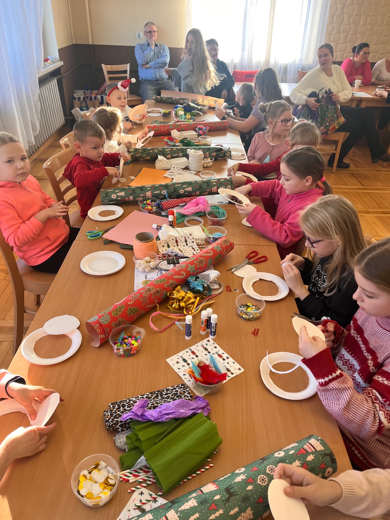 Grupa dzieci przy stole w sali, uczestnicząca w warsztatach plastycznych, tworząc ozdoby z papierowych talerzy, kolorowych koralików i bibuły, pod okiem dorosłych.