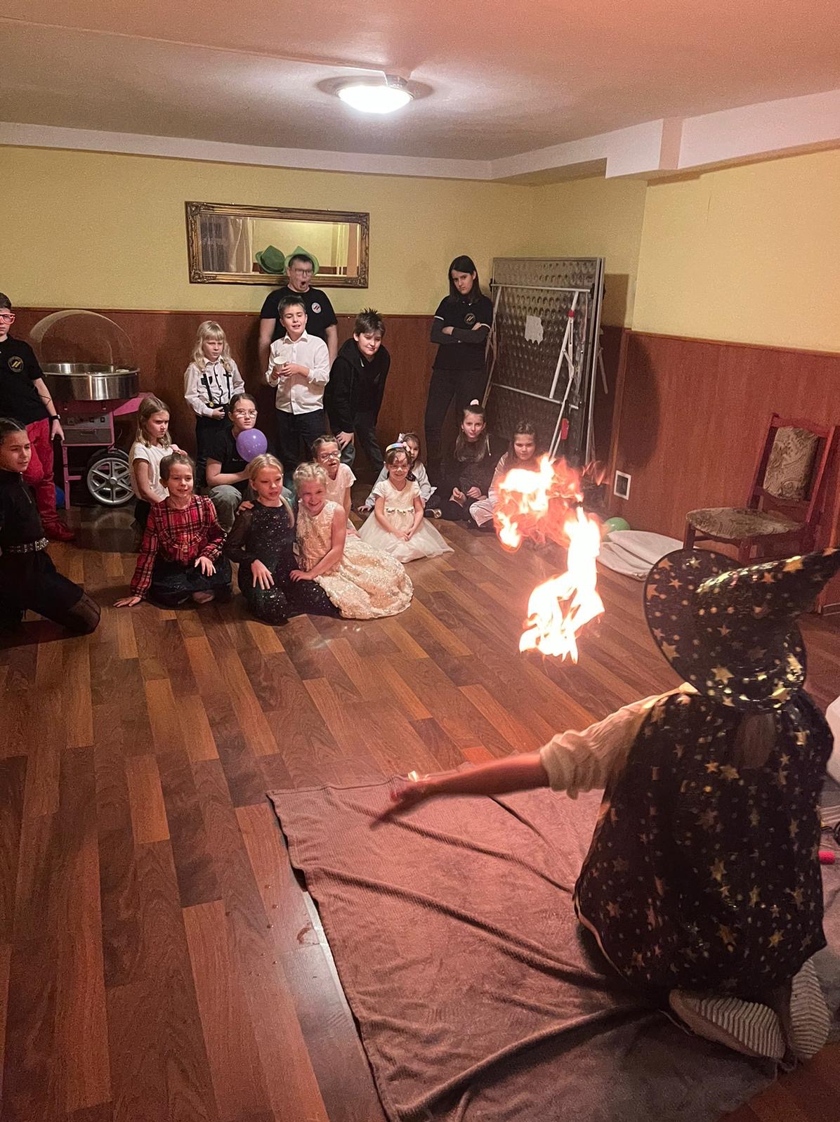 Osoba w stroju czarodzieja siedzi na podłodze, prezentując płomień przed grupą dzieci siedzących w rzędzie, z maszyną do waty cukrowej w tle po lewej stronie.