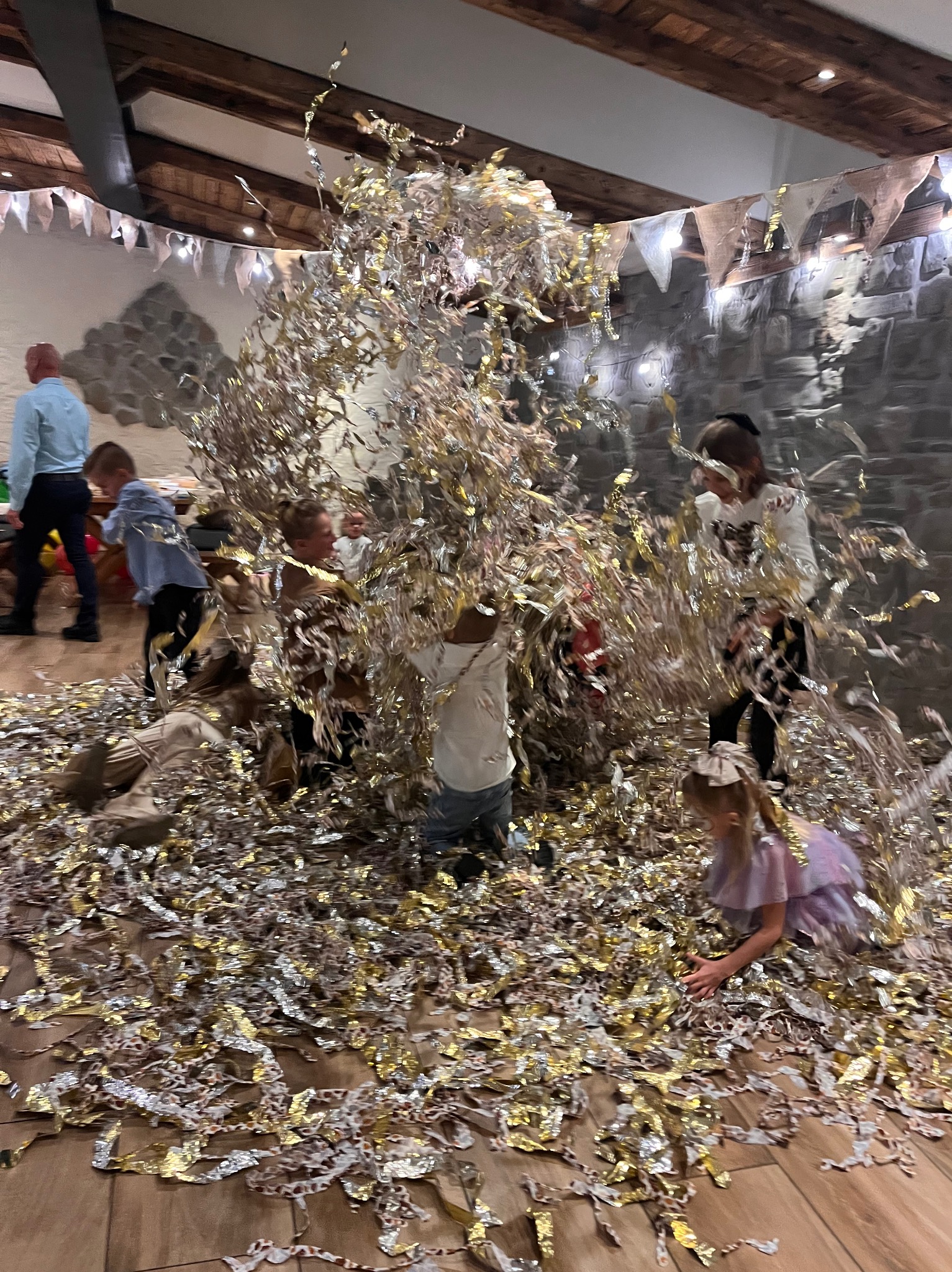 Dzieci bawią się w pomieszczeniu zasypanym złotym i srebrnym konfetti podczas imprezy, w tle kamienna ściana i dekoracyjne girlandy.