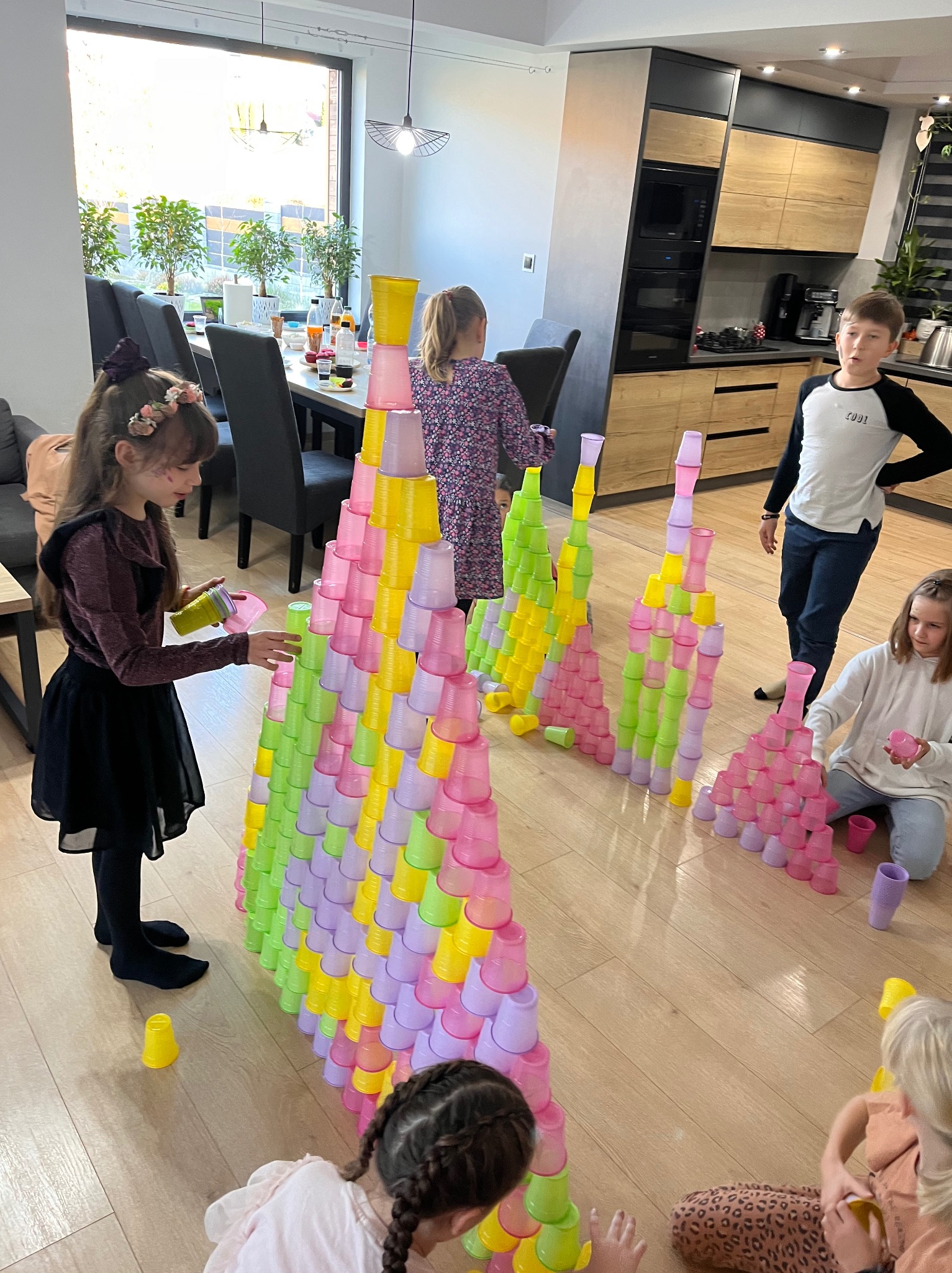 Dzieci budują piramidy z kolorowych plastikowych kubków w jasnym pokoju, widoczne różne etapy budowy, od pojedynczych kubków na podłodze po wysokie konstrukcje.