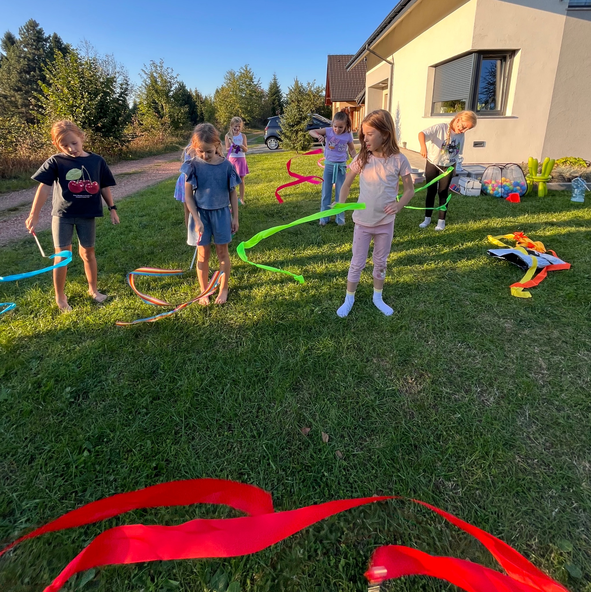 Grupa dzieci na trawie bawi się kolorowymi wstążkami, w tle dom i drzewa, widać akcesoria do zabawy.