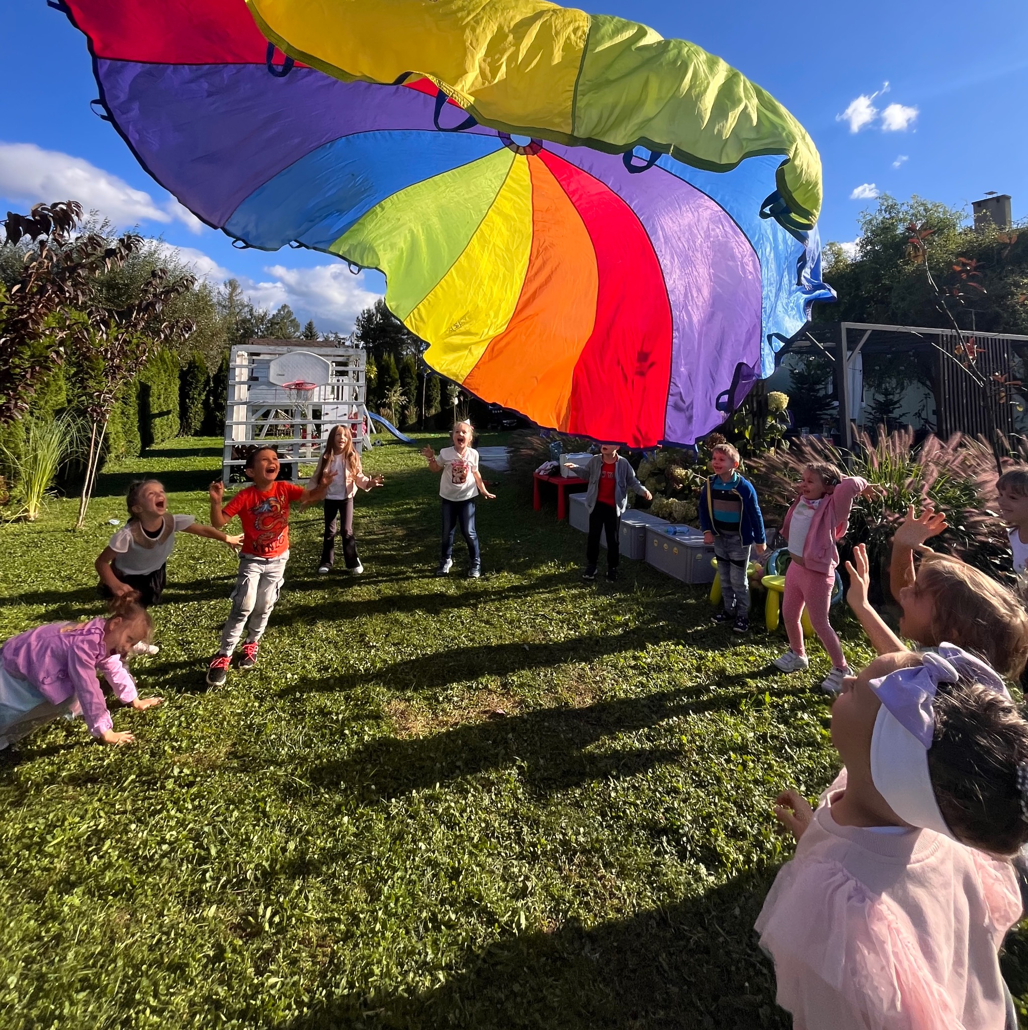 Grupa roześmianych dzieci bawi się na trawie pod dużym, kolorowym spadochronem w tęczowych barwach, w tle widoczny biały plac zabaw z koszem do koszykówki i zielone krzewy w słoneczny dzień.
