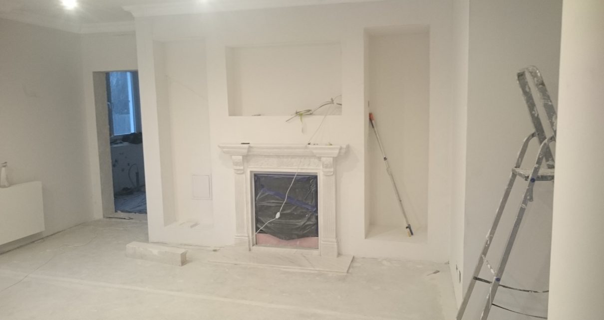 Wnętrze pokoju w trakcie malowania na biało, z zabezpieczonym kominkiem, widoczną drabiną i wałkiem do malowania.