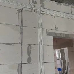 Białe rurki instalacyjne prowadzone po ścianie z betonu komórkowego i suficie w surowym stanie, z widocznymi zapiskami na ścianie, w tle otwór drzwiowy.