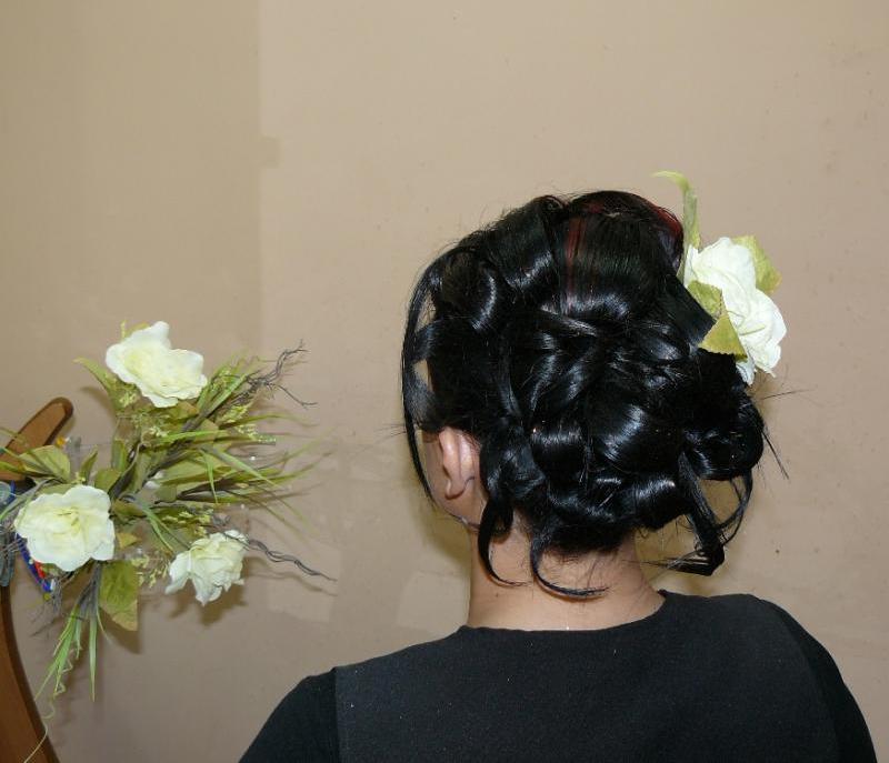 Upięcie włosów z ciemnych loków z wpiętym białym kwiatem, widok z tyłu, obok bukiet białych róż na stole.