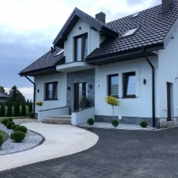 Elewacja jasnego domu jednorodzinnego z ciemną dachówką, ozdobnymi krzewami i dwukolorową ścieżką z kostki brukowej prowadzącą do wejścia.