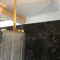 Deszczownica w kolorze złotym z włączonym strumieniem wody na tle ściany z czarnego marmuru z białymi żyłami, widoczny fragment sufitu z ozdobnym sztukaterią.