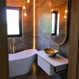 Elegancka łazienka z marmurowymi ścianami, wolnostojącą białą wanną, złotą armaturą i umywalką w kształcie misy, oświetlona wiszącymi żarówkami Edison i ledowym paskiem.